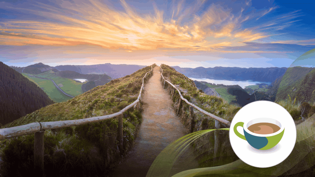 A scenic ridge path lined with wooden railings, overlooking lush green valleys, lakes, and mountains under a colourful sunset sky.
