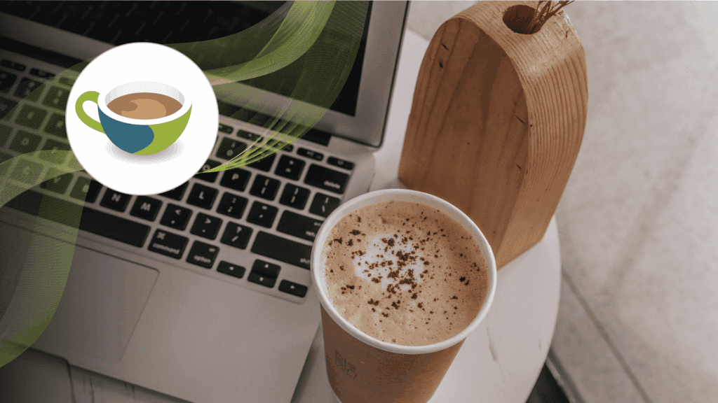 Laptop keyboard with a takeaway cup of frothy coffee on a small round table, alongside a wooden décor piece.